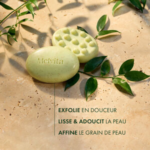 Savon Exfoliant Lissant L’Or Végétal 125 g, Deux savons sont au centre du visuel avec des plantes. Les deux côtés du savon sont visibles. En dessous, "Exfolie en douceur, Lisse et adoucit la peau, Affine le grain de peau" | Melvita