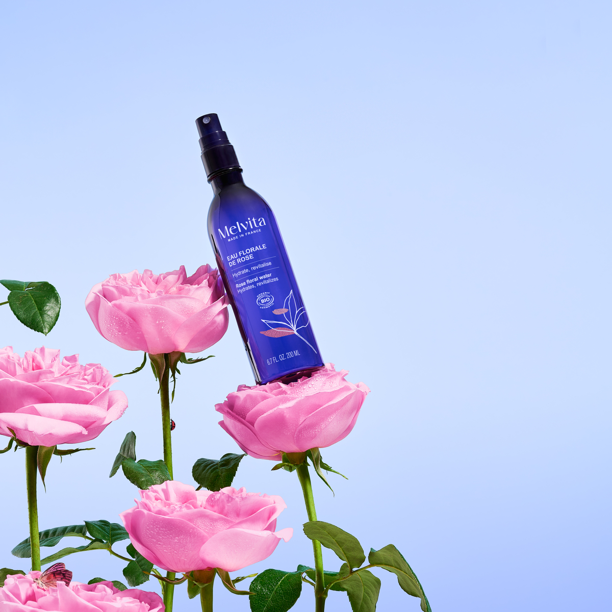 Eau Florale De Rose Bio en spray 200ml, Le produit tient en diagonale sur deux roses sur un fond bleu clair | Melvita